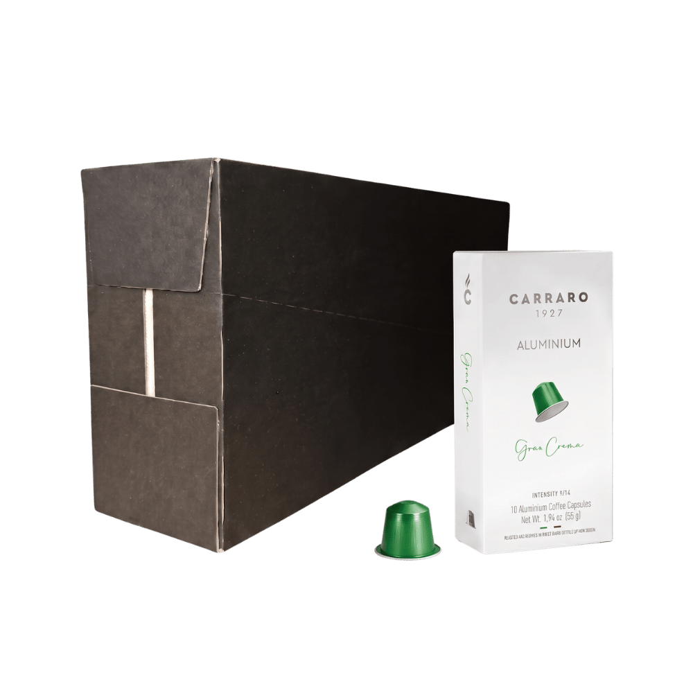 A large matte black rectangular box stands next to a small white package labeled CARRARO 1923 ALUMINIUM gran crema coffee capsules, with one green capsule placed in front on a clean white background.